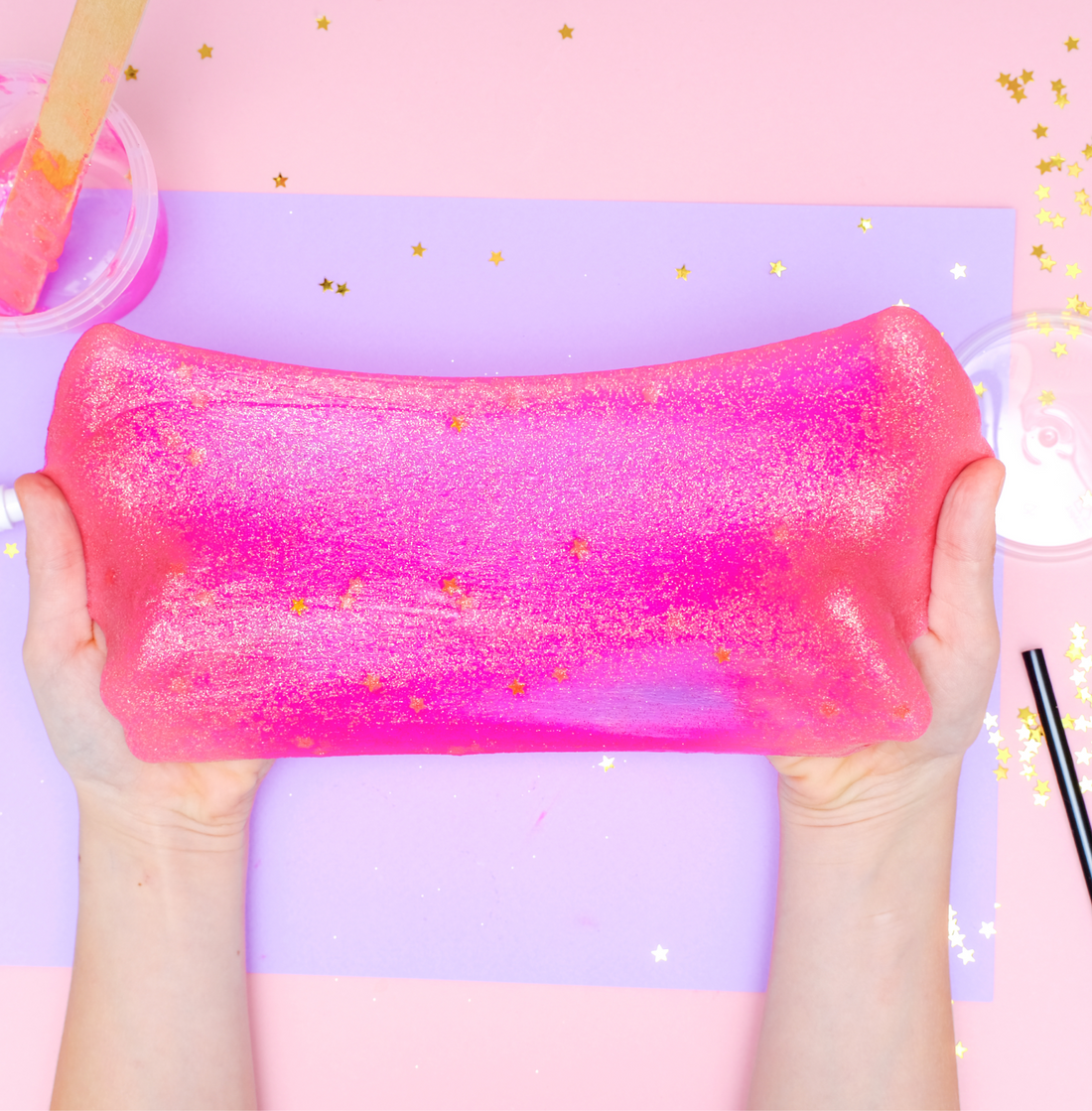 Hands stretching a wide sheet of glittery pink slime against a pastel pink and lilac background scattered with gold stars