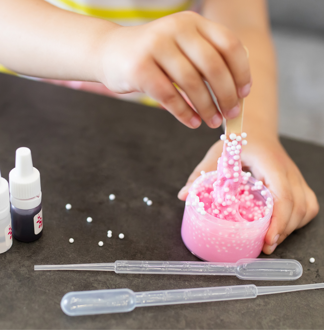 Child's hand sprinkling pink beads into slime on a dark surface, with a white dropper bottle of activator alongside