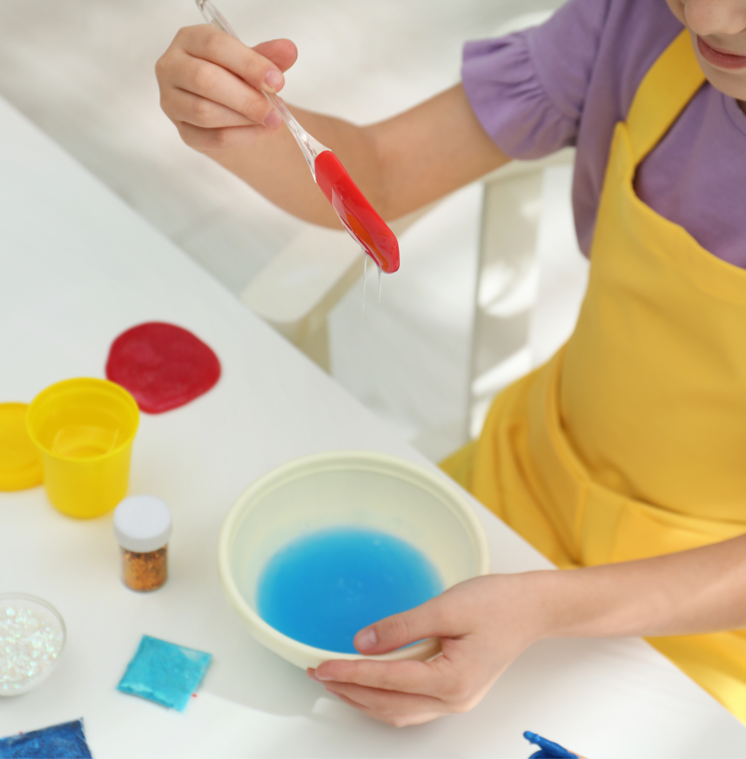 Child in a yellow apron using a red spatula to lift slime from a white surface, with red and yellow pots nearby