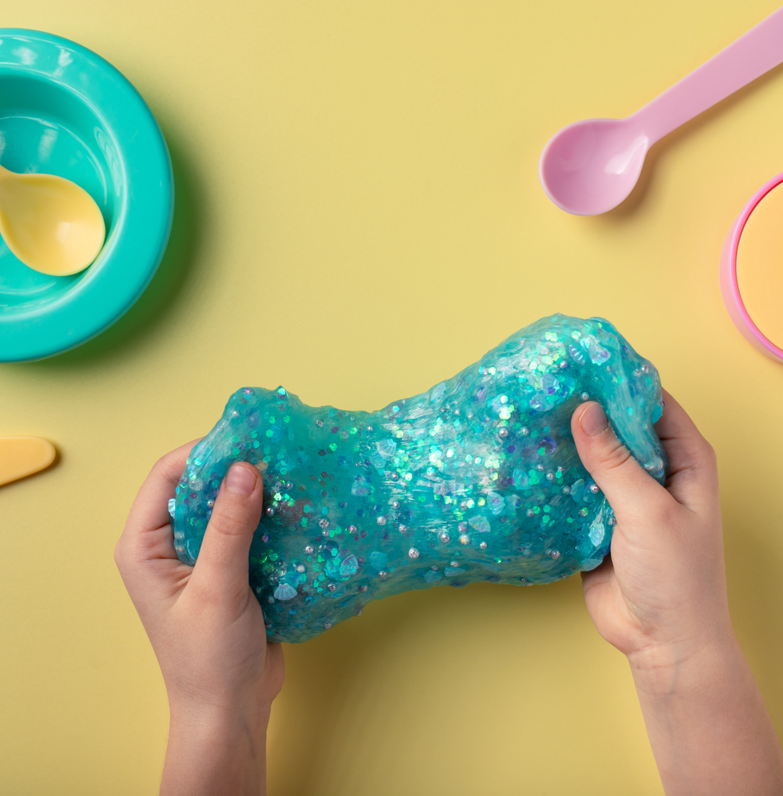 Hands pressing teal glitter slime on a yellow surface beside a teal bowl and a pink spoon