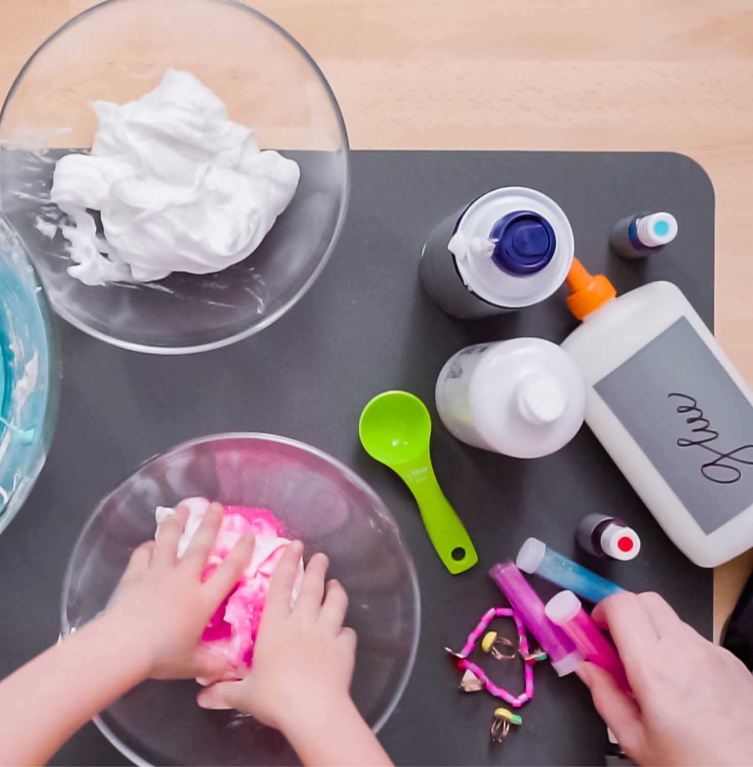 Slime experiment set-up: a glass bowl of white foam on a grey mat with glue bottles, a pigment dropper and a green measuring spoon