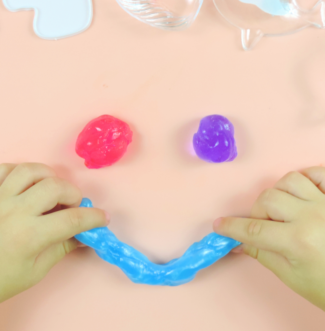Overhead view of pink and purple slime blobs on a peach surface beside clear storage containers, with a child's hands at the edge