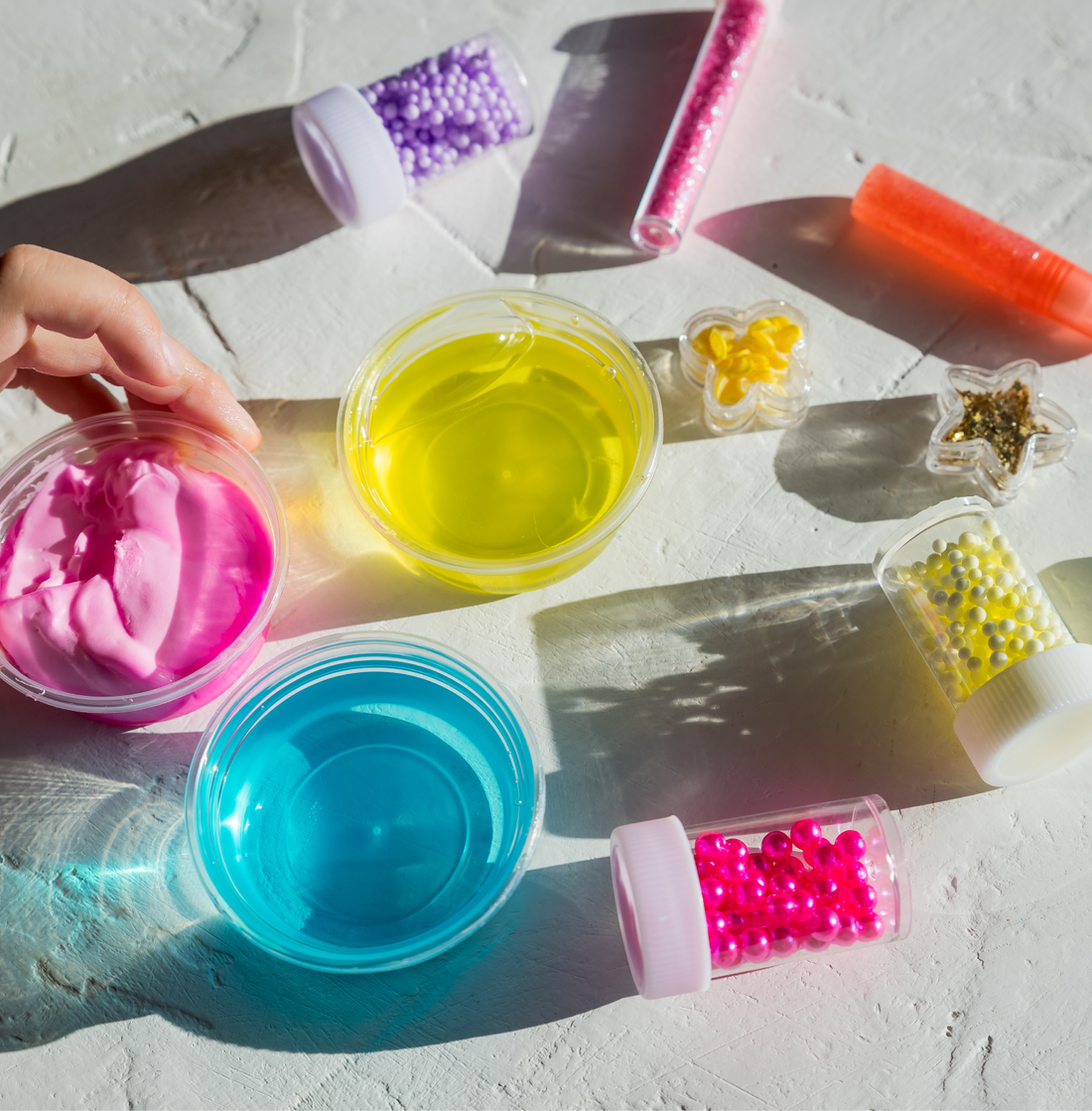 Slime-making components laid out in sunlight: tubs of pink, yellow and blue slime with pots of beads, glitter and activator bottles